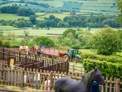 An image of a green tractor with a red trailer dropping off passengers from the tractor & trailer ride at Cantref Adventure Farm. There's a stunning backdrop of the Brecon countryside behind.